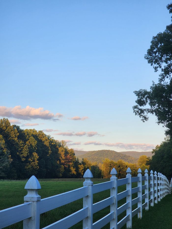 Scenic landscape with a white fence, lush greenery, and distant hills under a sunset sky.